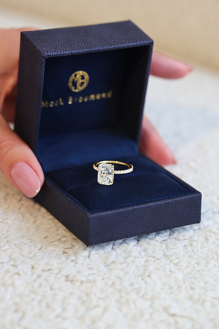 Woman holding a blue Mark Broumand ring box holding a yellow gold radiant cut engagement ring. 